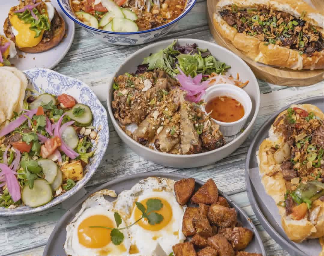 A generous catering spread of Bonjour Saigon dishes — bánh mì, noodle bowls, burgers, and Vietnamese breakfast plates.